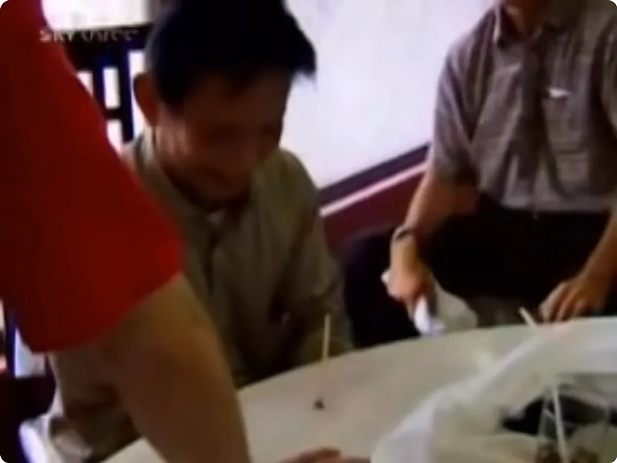 A broadcast still: a man leaning over a round table on which a chopstick stands upright, apparently driven into the tabletop, watched by two other men.