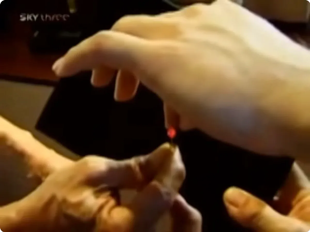 A broadcast still, close on two pairs of hands: a small LED held between fingertips is lit red, with no visible power source.