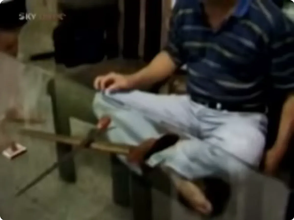 A broadcast still: a small sword resting across the slatted seat of a bench, hilt outward, beside a seated man in a striped shirt.