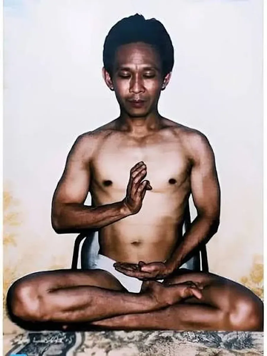 John Chang seated cross-legged in meditation, bare-chested, one hand raised palm-inward at the chest and the other resting open in his lap.