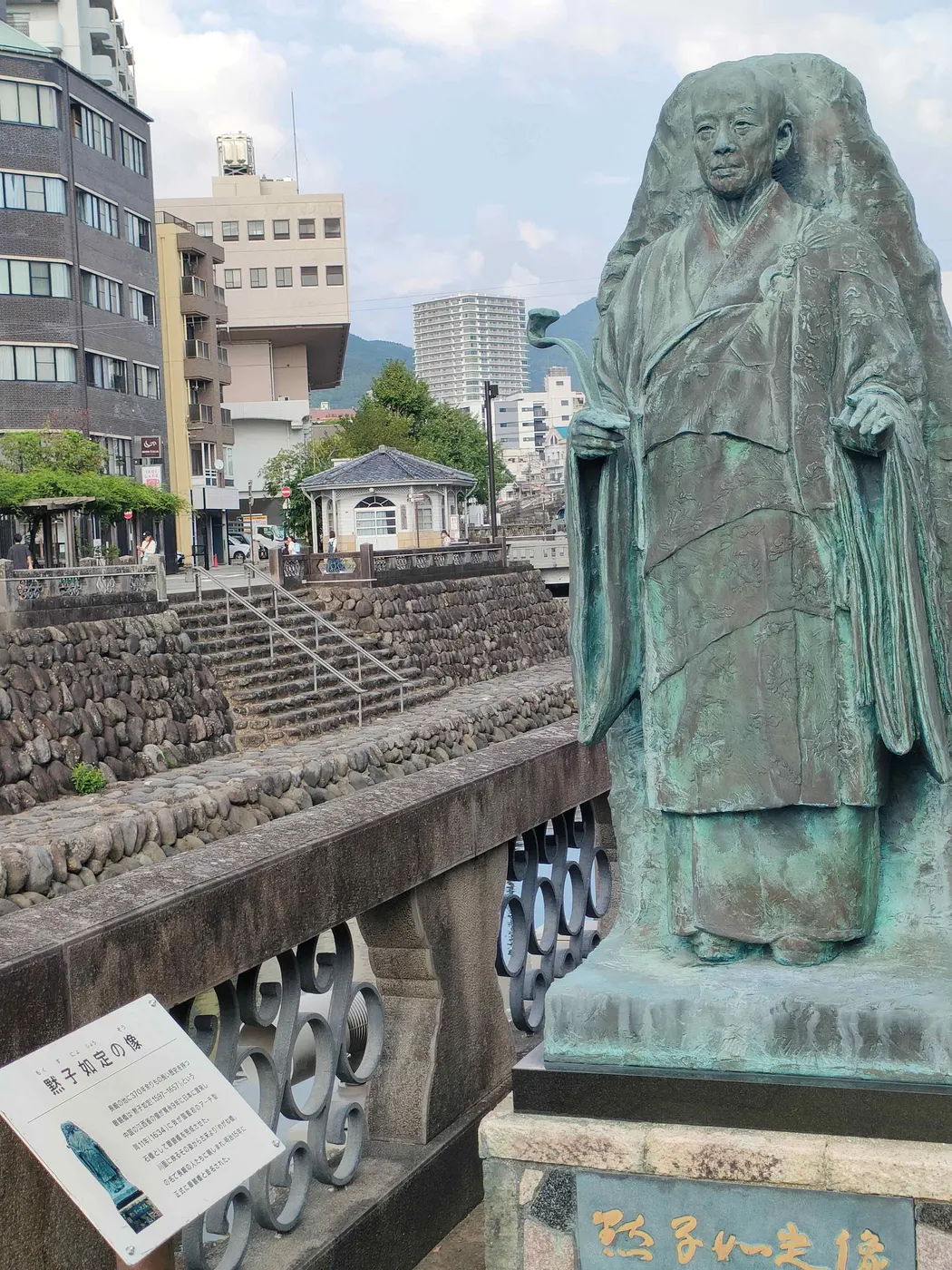 Modern stone statue of Mozi standing with a scroll, against a plain sky.